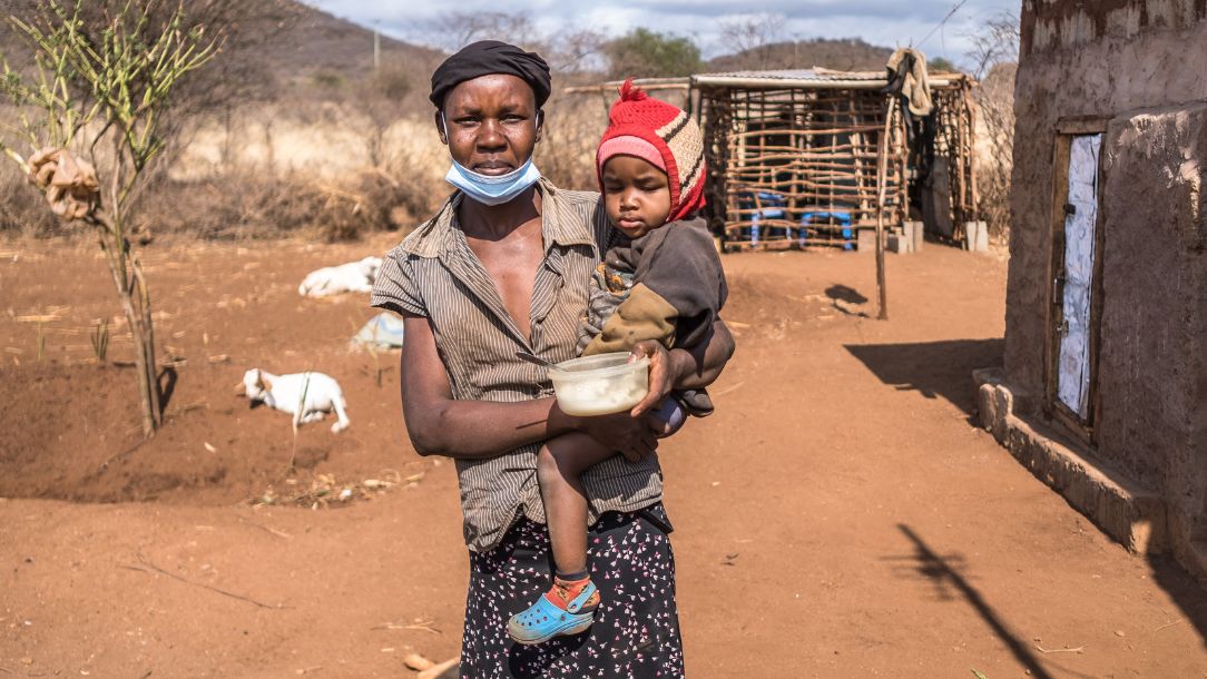 A mother holds her baby in a dusty landscape in Taita Taveta in Kenya. She is surrounded by starving animals.