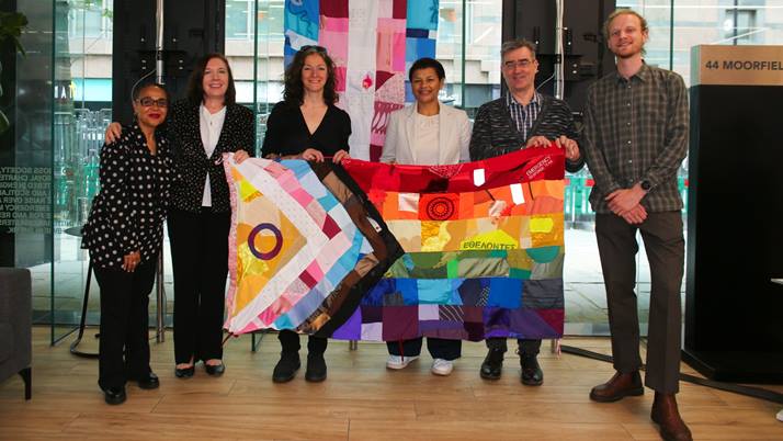 Four women and two men, including the British Red Cross CEO, hold the progress pride flag. They're stood in the UK head office in front of the trans flag