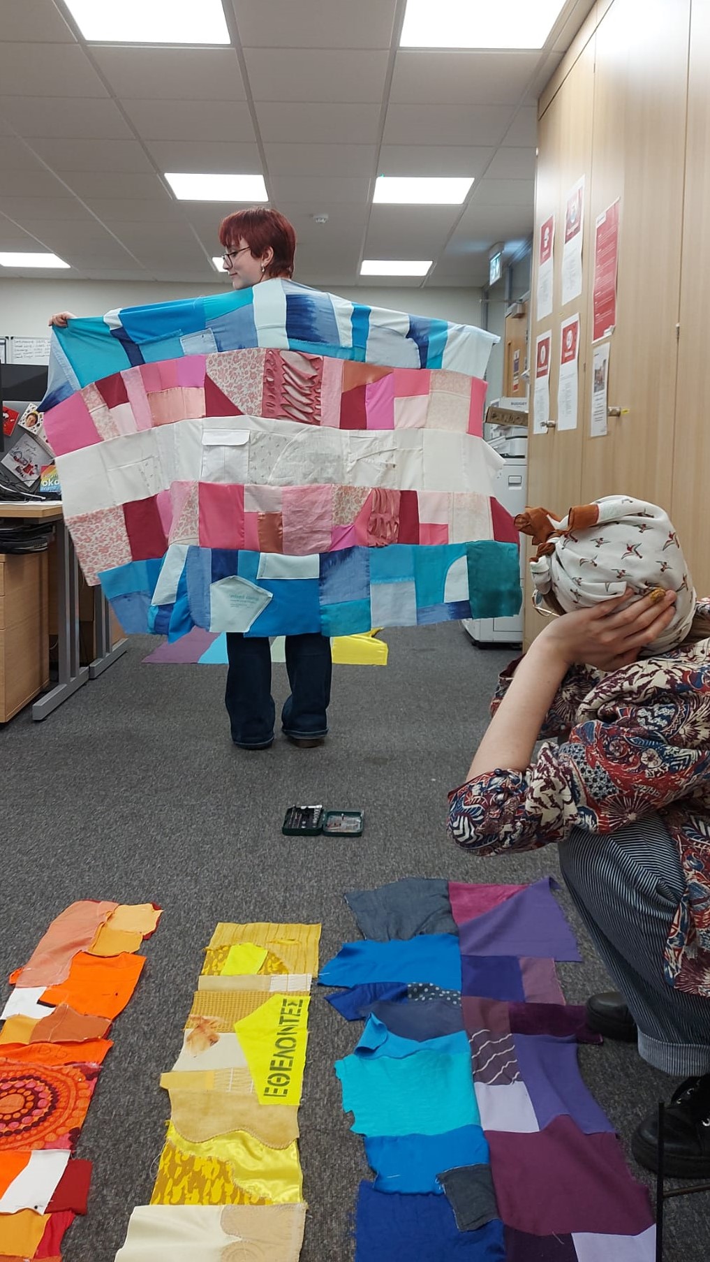 Person holding the trans flag with their back to the camera. A half made pride flag is on the floor of the office